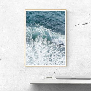 May include: A framed print depicting a seascape with a close-up of ocean waves. The artwork captures the dynamic movement of the water, with shades of teal and white creating depth and texture. The frame is a light wood colour.
