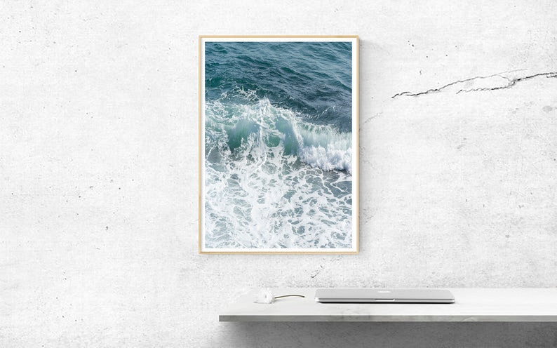 May include: A framed print depicting a seascape with a close-up of ocean waves. The artwork captures the dynamic movement of the water, with shades of teal and white creating depth and texture. The frame is a light wood colour.