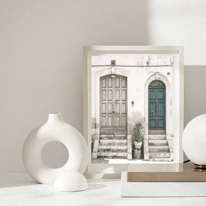 May include: A framed print of a white building with two doors. The door on the left is a weathered wood door with a paneled design. The door on the right is a solid teal blue door. A small potted plant sits on the steps in front of the doors.