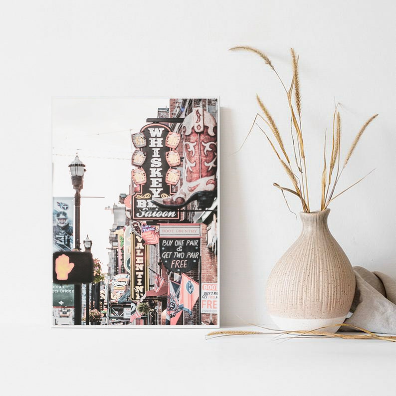 May include: A colorful photograph of a street scene in Nashville, Tennessee. The image features a variety of neon signs, including a large boot-shaped sign for a whiskey saloon. The street is lined with brick buildings and there are people walking around.
