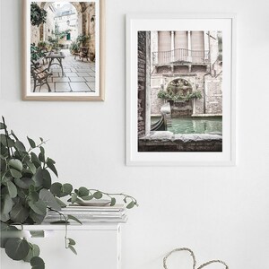 May include: Two framed art prints depicting Italian scenes. One shows a cafe with tables and chairs under an archway. The other features a canal with a gondola and a building with a balcony. The prints are in white frames.