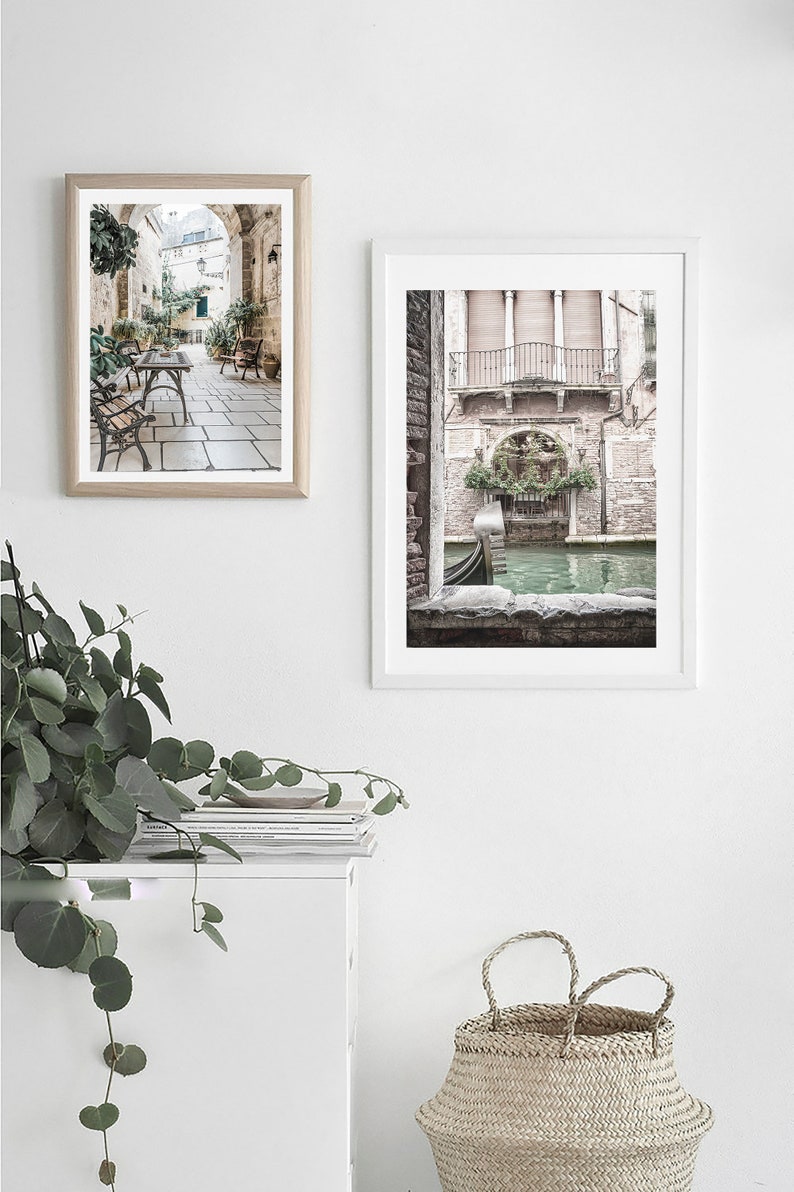 May include: Two framed art prints depicting Italian scenes. One shows a cafe with tables and chairs under an archway. The other features a canal with a gondola and a building with a balcony. The prints are in white frames.