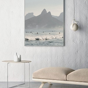 May include: A framed photograph of a beach scene with a mountain range in the background. The water is blue and there are waves crashing on the shore. There are people surfing in the water.