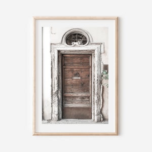 May include: A weathered wooden door with a metal ring handle, set in a white stone doorway with a decorative stone archway above. The door is framed by a light wood frame.