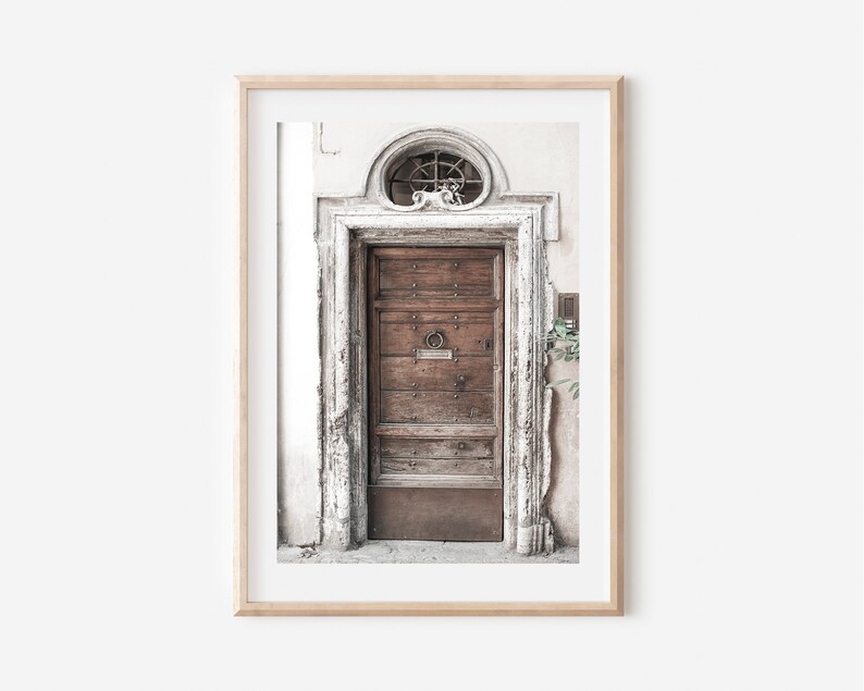 May include: A weathered wooden door with a metal ring handle, set in a white stone doorway with a decorative stone archway above. The door is framed by a light wood frame.