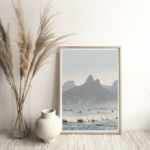 May include: A framed photograph of a beach scene with a mountain range in the background. The water is blue and there are waves crashing on the shore. There are people surfing in the water.