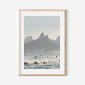 May include: A photograph of a beach scene with surfers in the water and mountains in the background. The sky is a light blue and the water is a deep blue. The mountains are a dark gray.