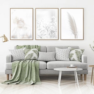 May include: Three framed minimalist art prints with a white background and subtle floral and feather designs. The prints are displayed on a white wall above a gray sofa with green throw pillows and a green throw blanket.