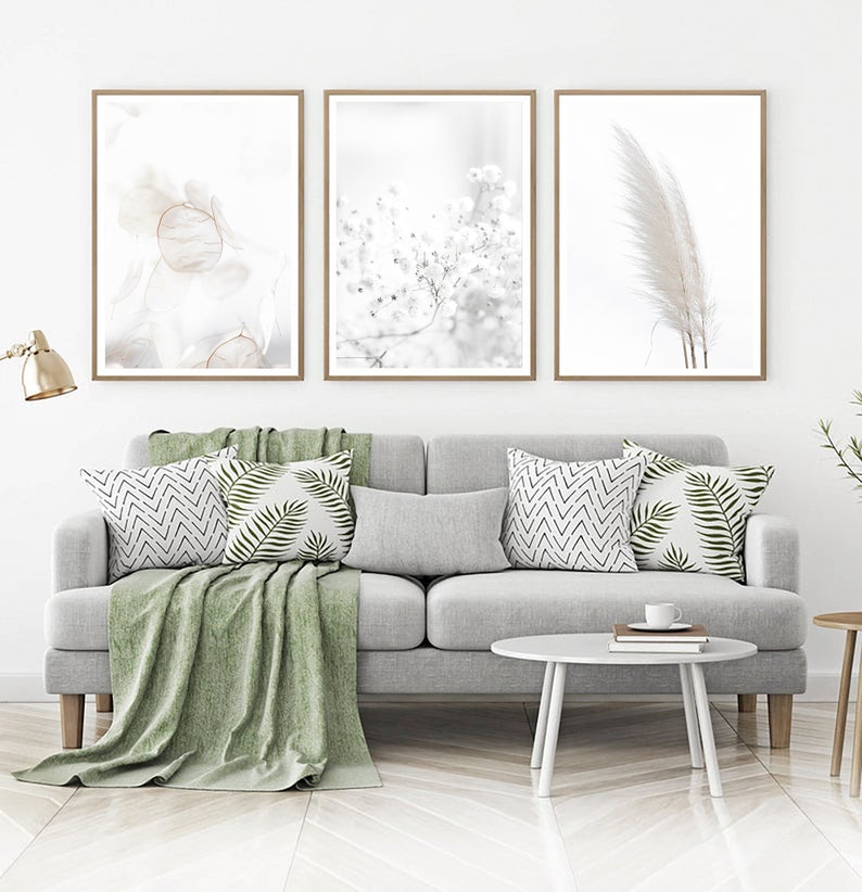 May include: Three framed minimalist art prints with a white background and subtle floral and feather designs. The prints are displayed on a white wall above a gray sofa with green throw pillows and a green throw blanket.