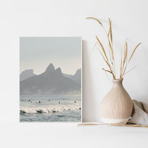 May include: A photograph of a beach scene with a mountain range in the background. The water is a light blue color and there are several people surfing in the waves. The sky is a light gray color.