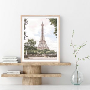 May include: A framed photograph of the Eiffel Tower in Paris, France. The tower is a light brown color and is surrounded by trees and greenery. The photo is in a light wood frame and is hanging on a white wall.