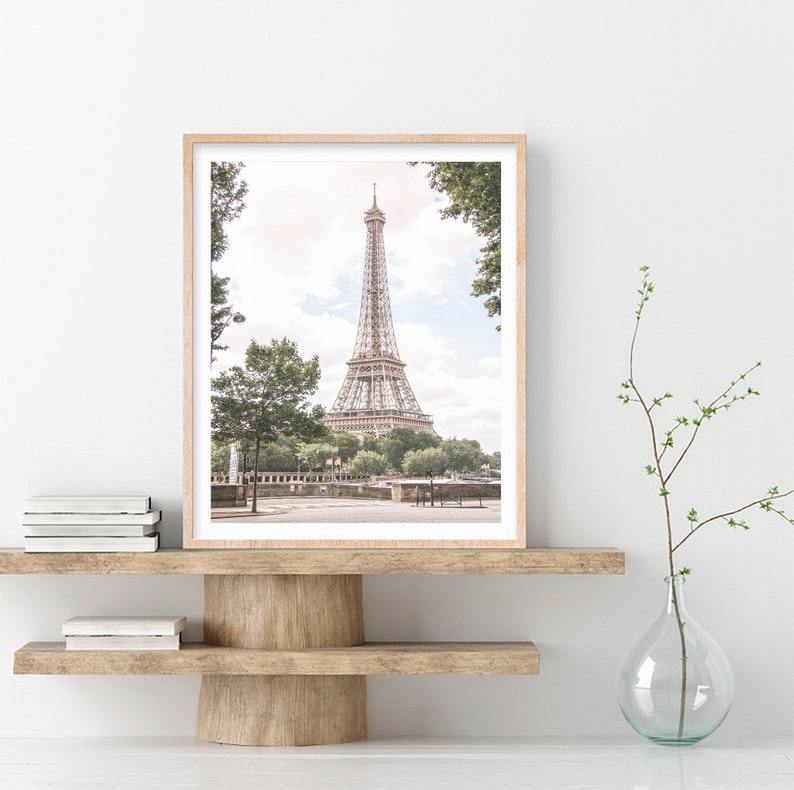 May include: A framed photograph of the Eiffel Tower in Paris, France. The tower is a light brown color and is surrounded by trees and greenery. The photo is in a light wood frame and is hanging on a white wall.