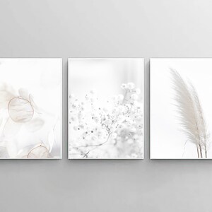 May include: Three minimalist art prints with a white background. The left print features a delicate, translucent, light brown floral design. The center print features a cluster of small white flowers. The right print features a single, long, white feather.