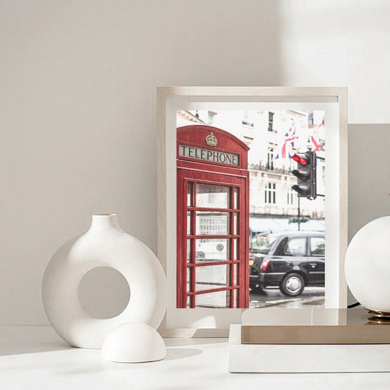 May include: A white ceramic vase with a circular opening in the middle, a white ceramic sphere, and a framed photograph of a red telephone booth in London, England. The photograph also shows a black taxi cab and a traffic light.