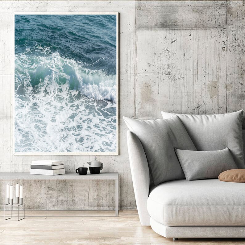 May include: A framed print showing a close-up of ocean waves. The image captures the movement of the water, with shades of teal and white. The artwork is displayed on a concrete wall above a grey table and white sofa.