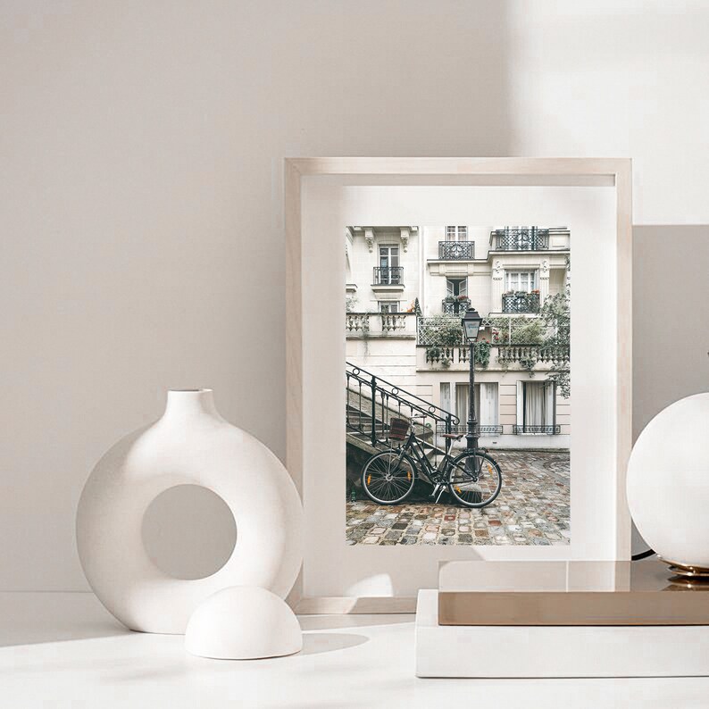 May include: A framed photograph of a Parisian street scene with a bicycle parked on a cobblestone street. The building is white with black accents and has a wrought iron balcony. The photo is in a white frame and is sitting on a white surface.