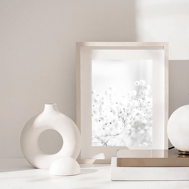 Pode incluir: Um vaso de cer&acirc;mica branco com uma abertura circular no centro, uma esfera de cer&acirc;mica branca e uma fotografia emoldurada de um arranjo de flores brancas. A moldura &eacute; de madeira clara e a foto est&aacute; em preto e branco.