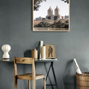 May include: A framed photograph of a city skyline with tall buildings and trees in the foreground. The photo is hanging on a gray wall above a desk with a wooden chair and a lamp.