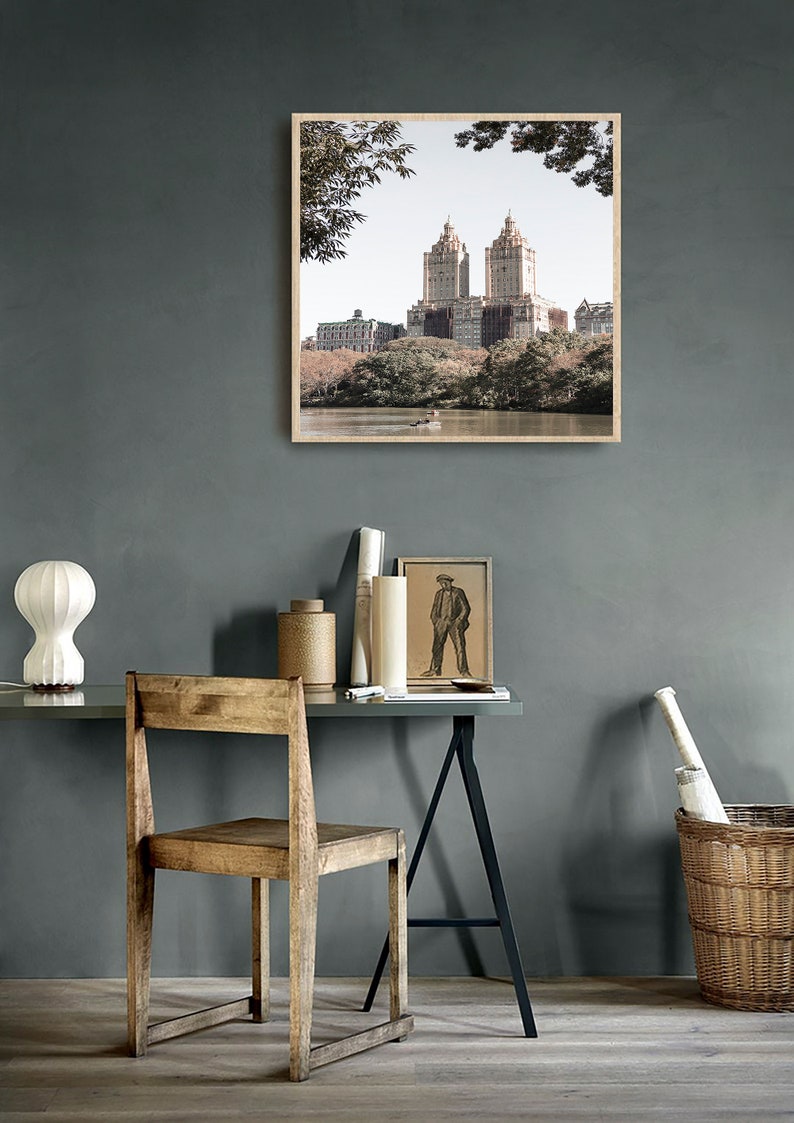 May include: A framed photograph of a city skyline with tall buildings and trees in the foreground. The photo is hanging on a gray wall above a desk with a wooden chair and a lamp.