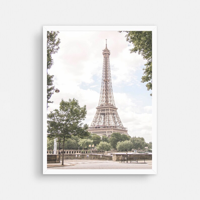 May include: A view of the Eiffel Tower in Paris, France. The tower is seen from a distance, with trees and a bridge in the foreground. The sky is a light blue with some clouds.