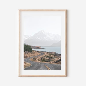 May include: A winding road leads through a mountain landscape with a lake in the distance. The mountains are snow-capped and the sky is a light blue.
