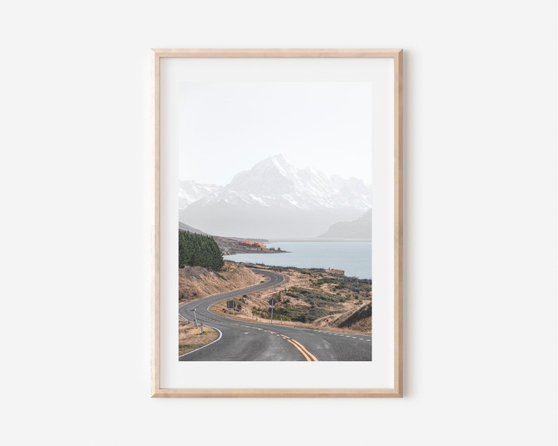 May include: A winding road leads through a mountain landscape with a lake in the distance. The mountains are snow-capped and the sky is a light blue.