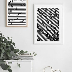 May include: Two black and white framed prints of people walking on a crosswalk with diagonal stripes. The prints are hanging on a white wall above a white shelf with a stack of books and a woven basket.