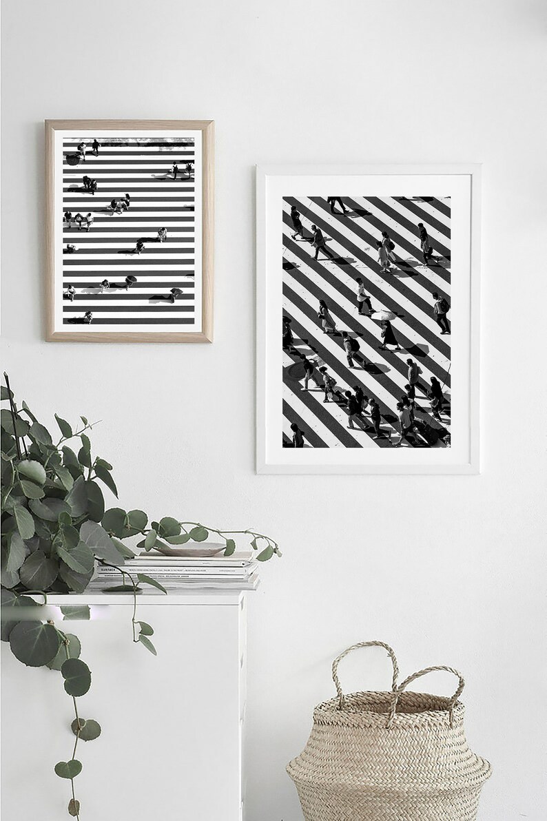 May include: Two black and white framed prints of people walking on a crosswalk with diagonal stripes. The prints are hanging on a white wall above a white shelf with a stack of books and a woven basket.