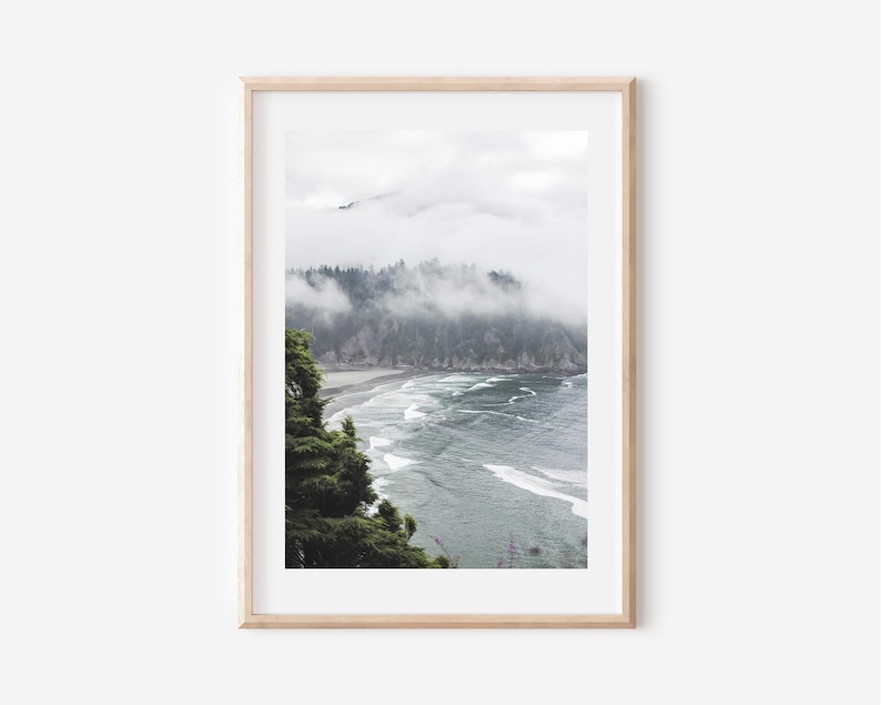 May include: A framed print of a coastal landscape. The image features a beach with waves, a forested cliff, and a cloudy sky. The frame is a light wood color, and the overall aesthetic is muted and natural.