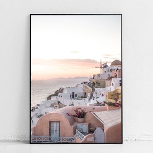 May include: A photograph of a white and pink village on a cliff overlooking the ocean. The sky is a soft pink and orange, and there are white buildings with blue accents. There is a windmill in the distance.