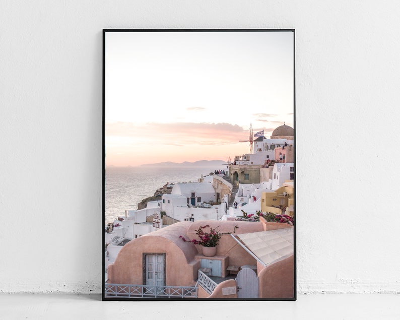 May include: A photograph of a white and pink village on a cliff overlooking the ocean. The sky is a soft pink and orange, and there are white buildings with blue accents. There is a windmill in the distance.