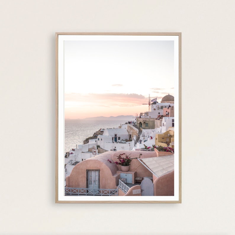 May include: A photograph of a white-washed village perched on a cliff overlooking the ocean. The sky is a soft pink and orange, and the sun is setting in the distance. The village is made up of small, white houses with blue doors and windows. There are also some windmills in the distance.