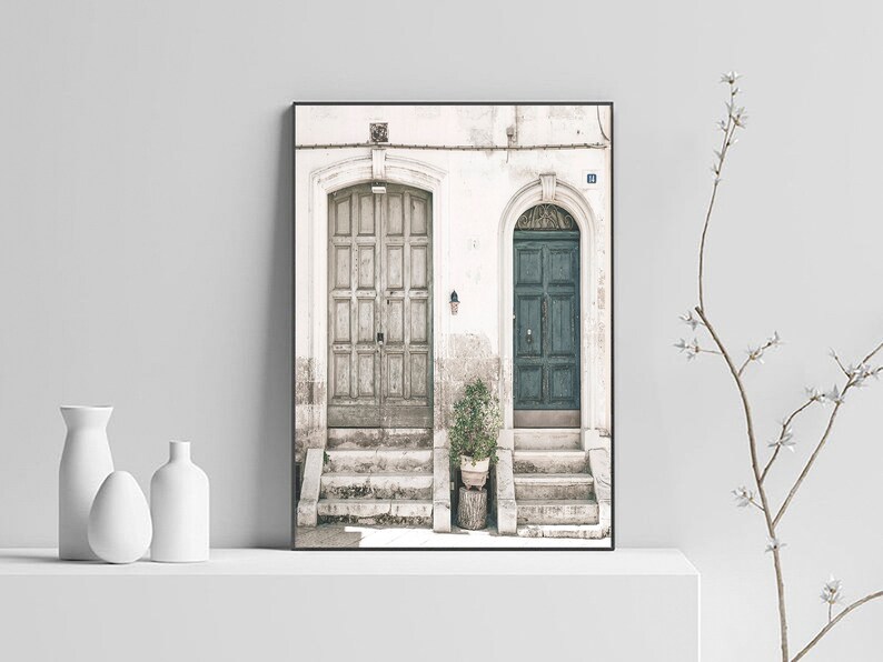 May include: A framed photograph of two doors, one painted a light brown and the other a dark green, with steps leading up to them. The doors are set against a white wall with a potted plant in front of the green door.