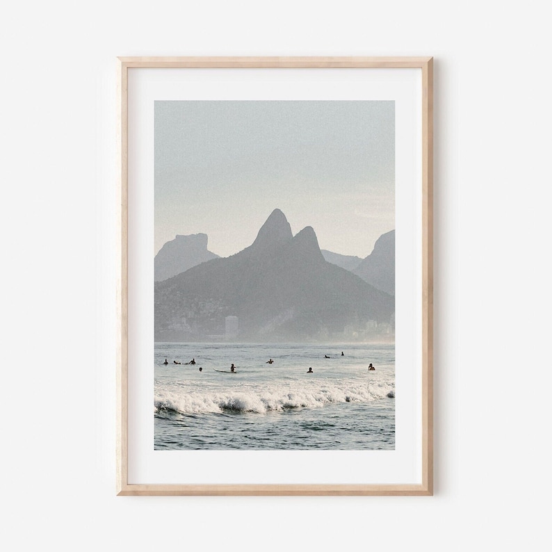 May include: A photograph of a beach scene with a mountain range in the background. The water is blue and there are waves crashing on the shore. There are people surfing in the water.