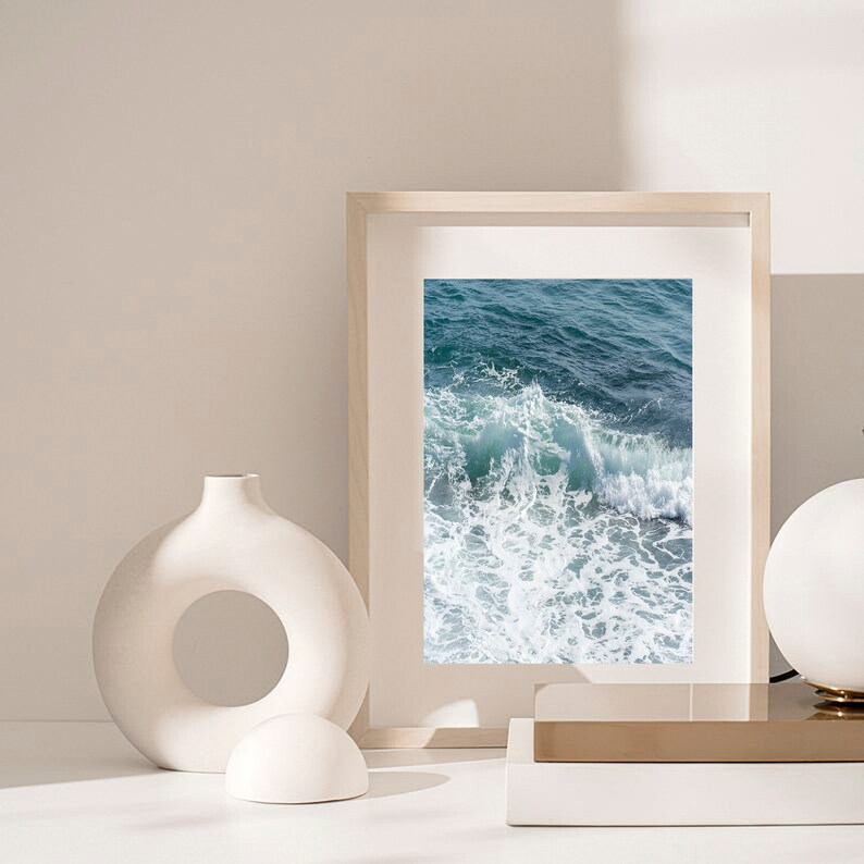 May include: A framed print showing a close-up of ocean waves. The image captures the turquoise and white of the water as it crashes. The frame is light-coloured, and the print is displayed on a white surface.