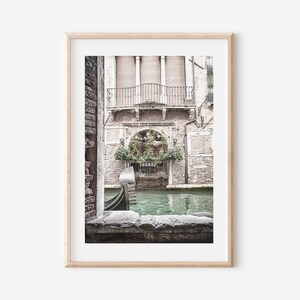 May include: Framed print of a Venetian canal scene. The image features a gondola on the water, framed by a stone archway. Buildings with balconies and lush greenery are visible in the background. The color palette is muted with soft tones.