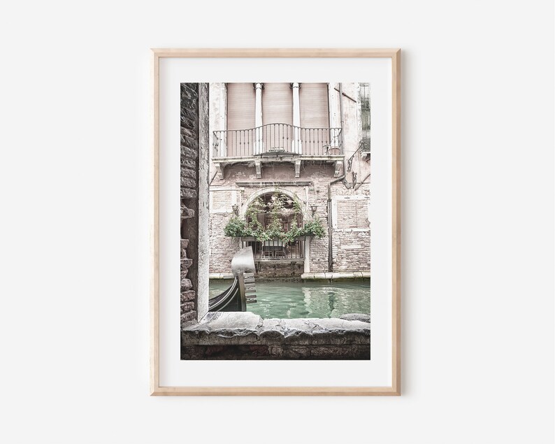 May include: Framed print of a Venetian canal scene. The image features a gondola on the water, framed by a stone archway. Buildings with balconies and lush greenery are visible in the background. The color palette is muted with soft tones.
