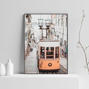 May include: A vintage yellow and white tram travels up a cobblestone street in a European city. The tram has large windows and a wooden frame. The street is lined with white buildings.