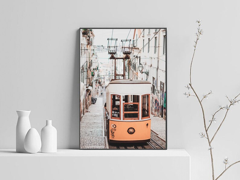May include: A vintage yellow and white tram travels up a cobblestone street in a European city. The tram has large windows and a wooden frame. The street is lined with white buildings.