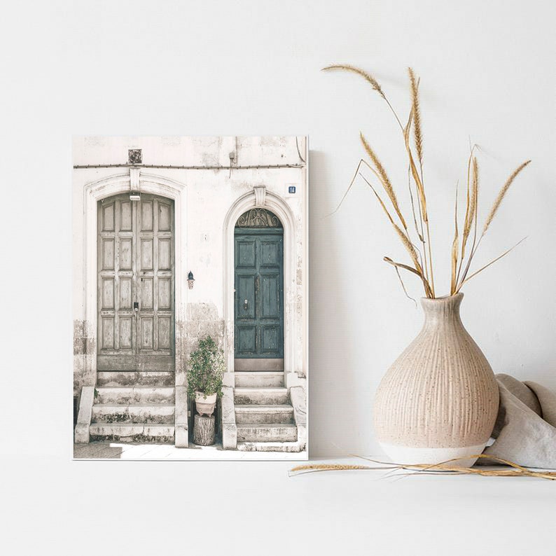 May include: A photograph of two doors, one painted a light green and the other a faded white, with steps leading up to them. The doors are set in a white building with a potted plant in front of the green door.