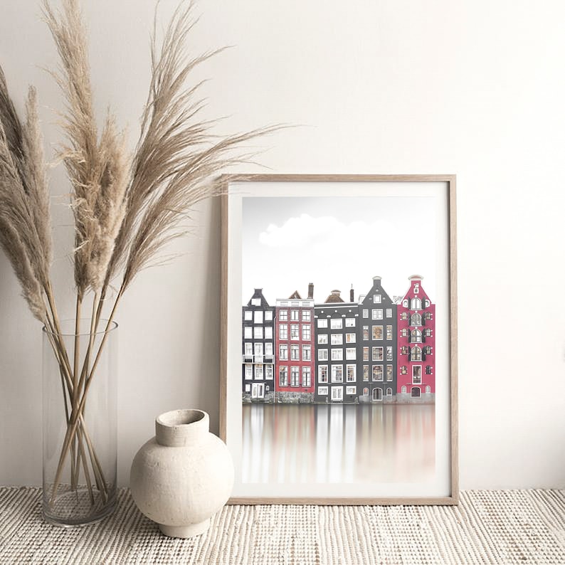 May include: A framed print of a row of colorful houses reflected in a canal. The houses are a mix of red, black, and white with white windows. The print has a light gray background.