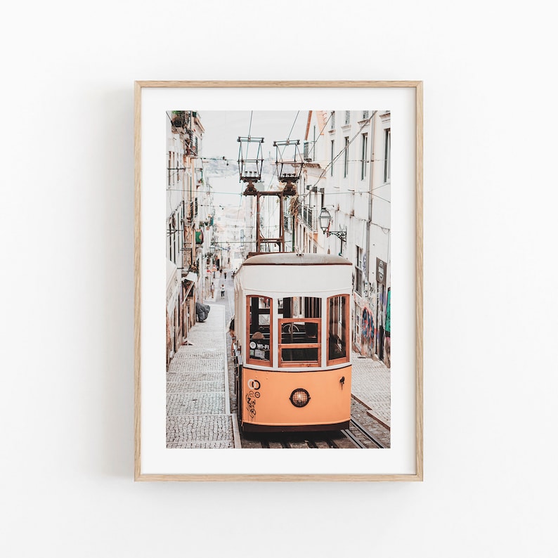 May include: A yellow and white vintage tram travels up a cobblestone street in Lisbon, Portugal. The tram is surrounded by buildings with colorful graffiti.