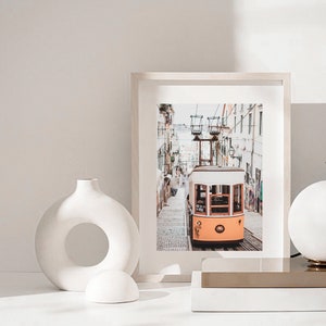 May include: A white ceramic vase with a circular opening in the middle, a white ceramic sphere, and a framed photograph of a yellow tram traveling up a hill in a city. The photograph is in a white frame with a white mat.