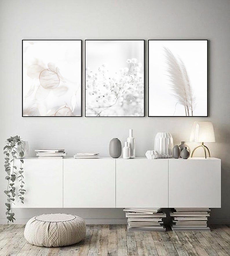 May include: Three minimalist black framed prints with white backgrounds and delicate floral and feather imagery. The prints are hung on a white wall above a white cabinet with a white countertop and a stack of books on either side.