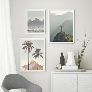 May include: Three framed prints of landscapes in Brazil. The first print shows a beach with surfers and mountains in the background. The second print shows a mountain with a statue of Christ the Redeemer on top. The third print shows two palm trees in front of a mountain.