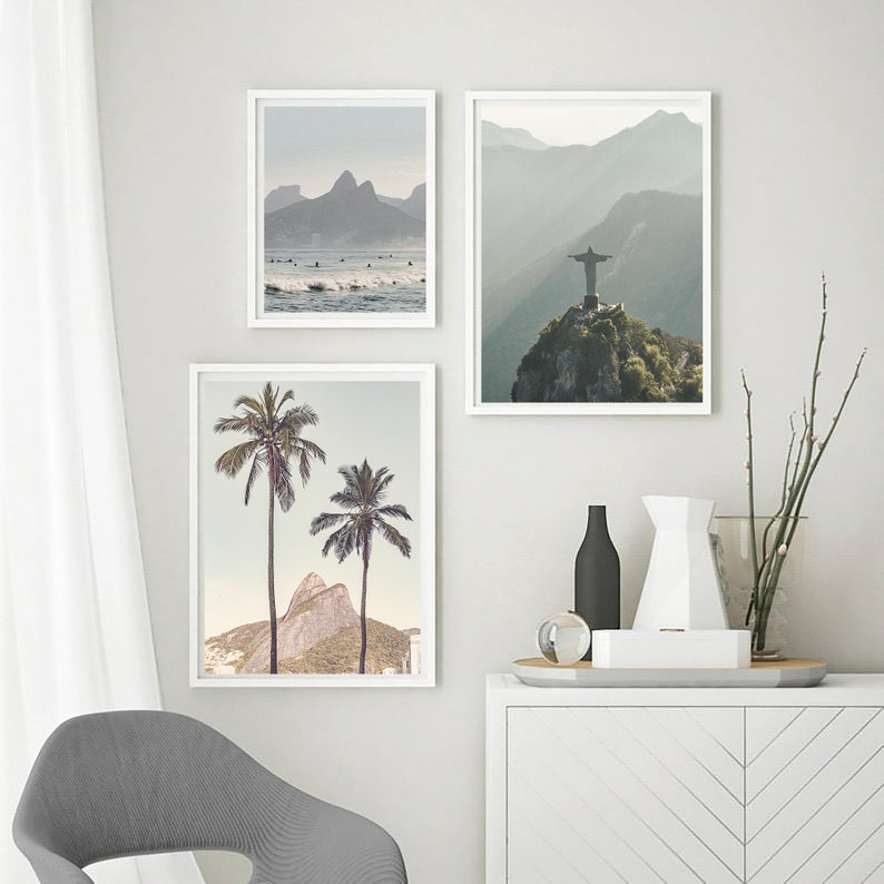 May include: Three framed prints of landscapes in Brazil. The first print shows a beach with surfers and mountains in the background. The second print shows a mountain with a statue of Christ the Redeemer on top. The third print shows two palm trees in front of a mountain.