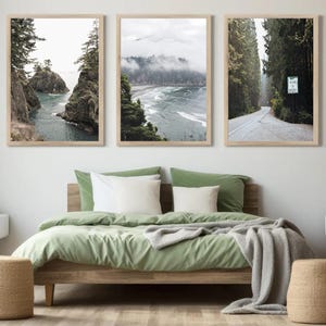 May include: Three framed landscape prints above a bed with green bedding and white pillows. The prints depict coastal scenes and a road through a forest. Two woven stools and two white nightstands with wooden legs complete the bedroom decor.