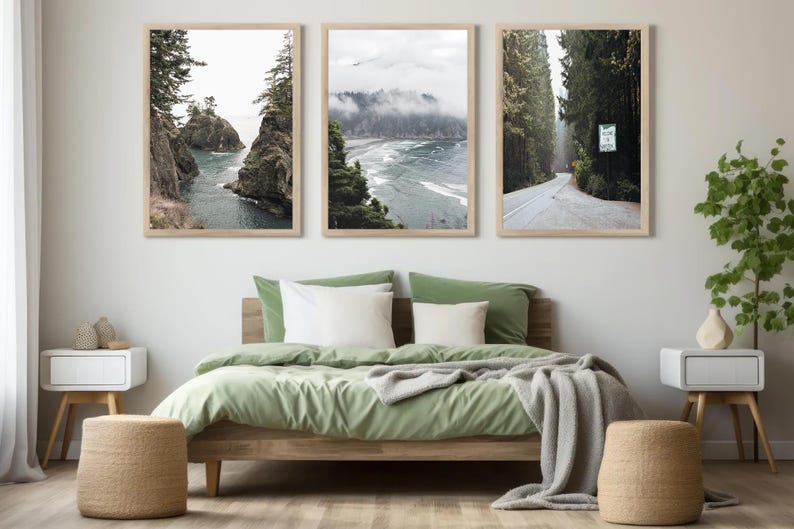 May include: Three framed landscape prints above a bed with green bedding and white pillows. The prints depict coastal scenes and a road through a forest. Two woven stools and two white nightstands with wooden legs complete the bedroom decor.