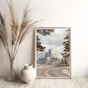 May include: A framed landscape print featuring a winding road with yellow lines leading towards a large mountain. The print is in a light wooden frame. Tall, dried grasses are in a clear glass vase, and a white ceramic vase sits nearby.