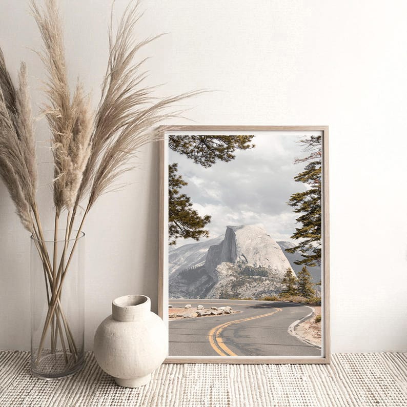 May include: A framed landscape print featuring a winding road with yellow lines leading towards a large mountain. The print is in a light wooden frame. Tall, dried grasses are in a clear glass vase, and a white ceramic vase sits nearby.